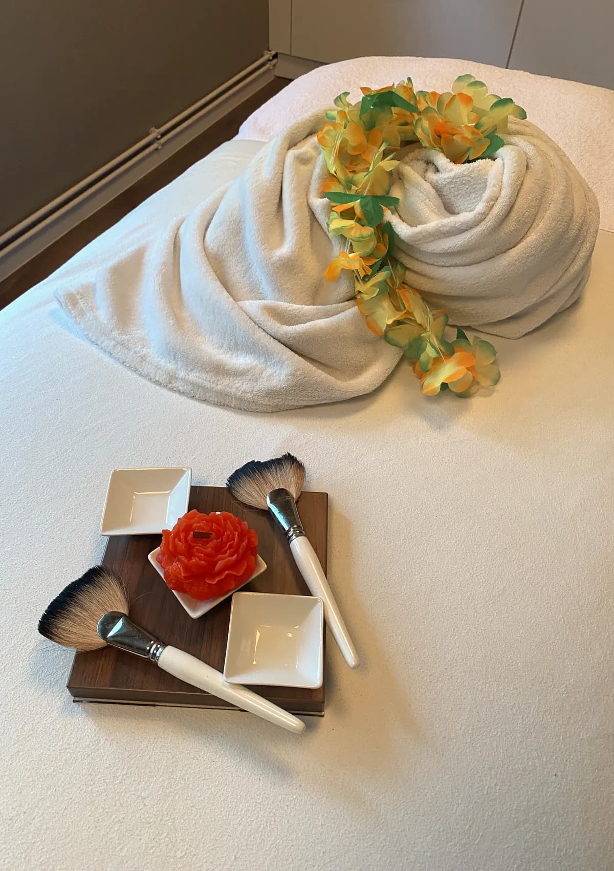 Behandlungsliege im Beauty Center Oberlausitz mit Holzbrett, Fächerpinseln, Keramikschälchen und roter Blütenkerze, dahinter ein Handtuch mit hawaiianischem Blütenkranz
